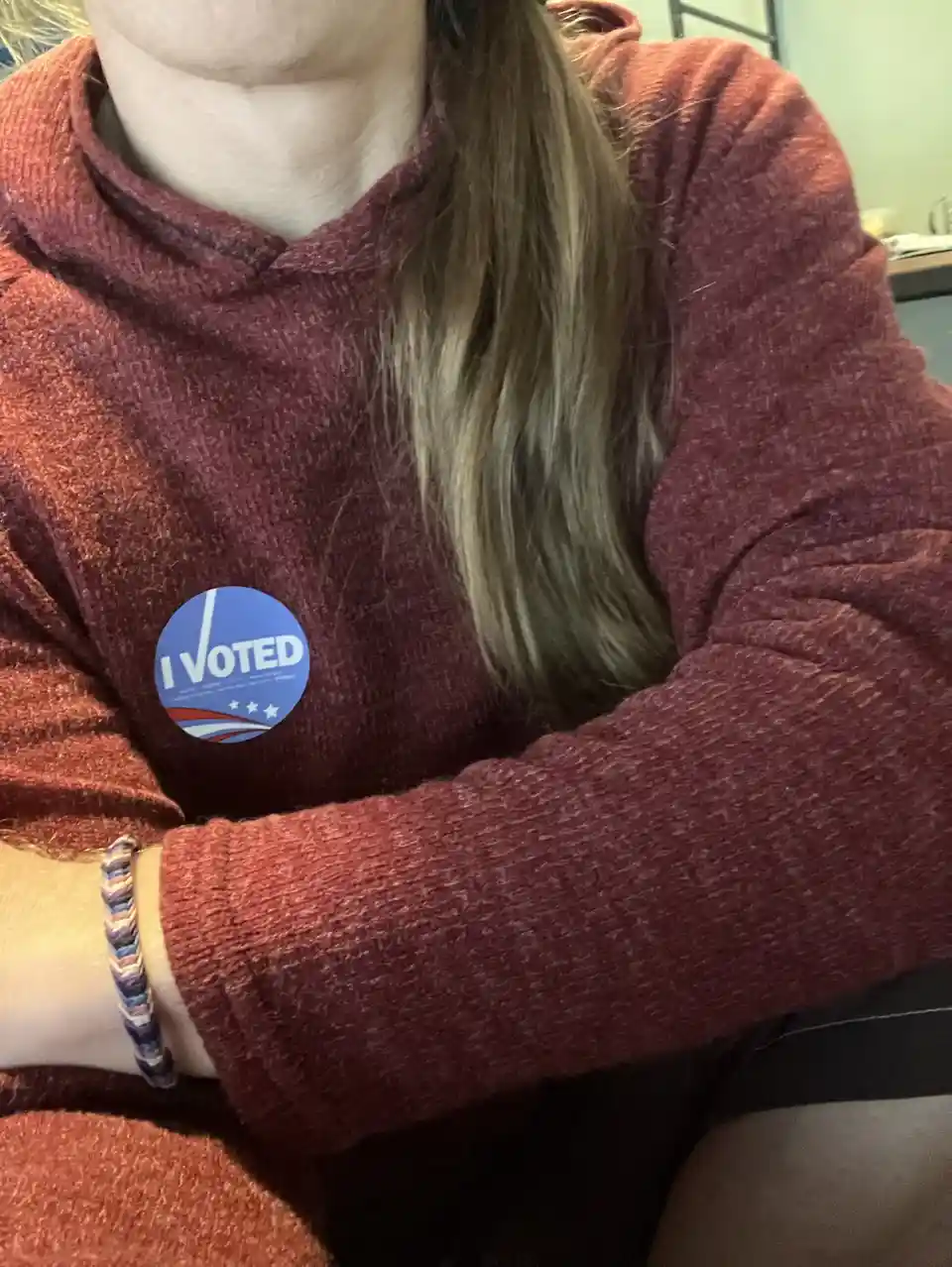 A close-up photo shows a person wearing a maroon hoodie with an 'I VOTED' sticker on the left chest. The sticker is blue…