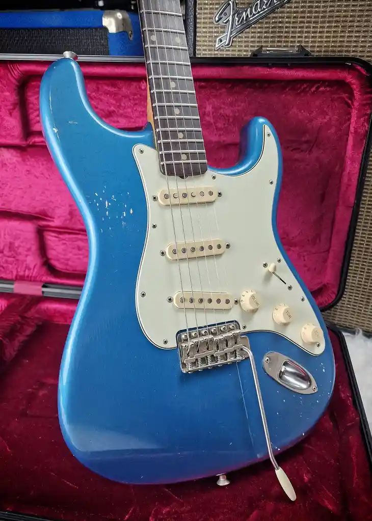 A blue Fender Stratocaster electric guitar with a white pickguard and cream knobs rests inside an open case lined with pink…