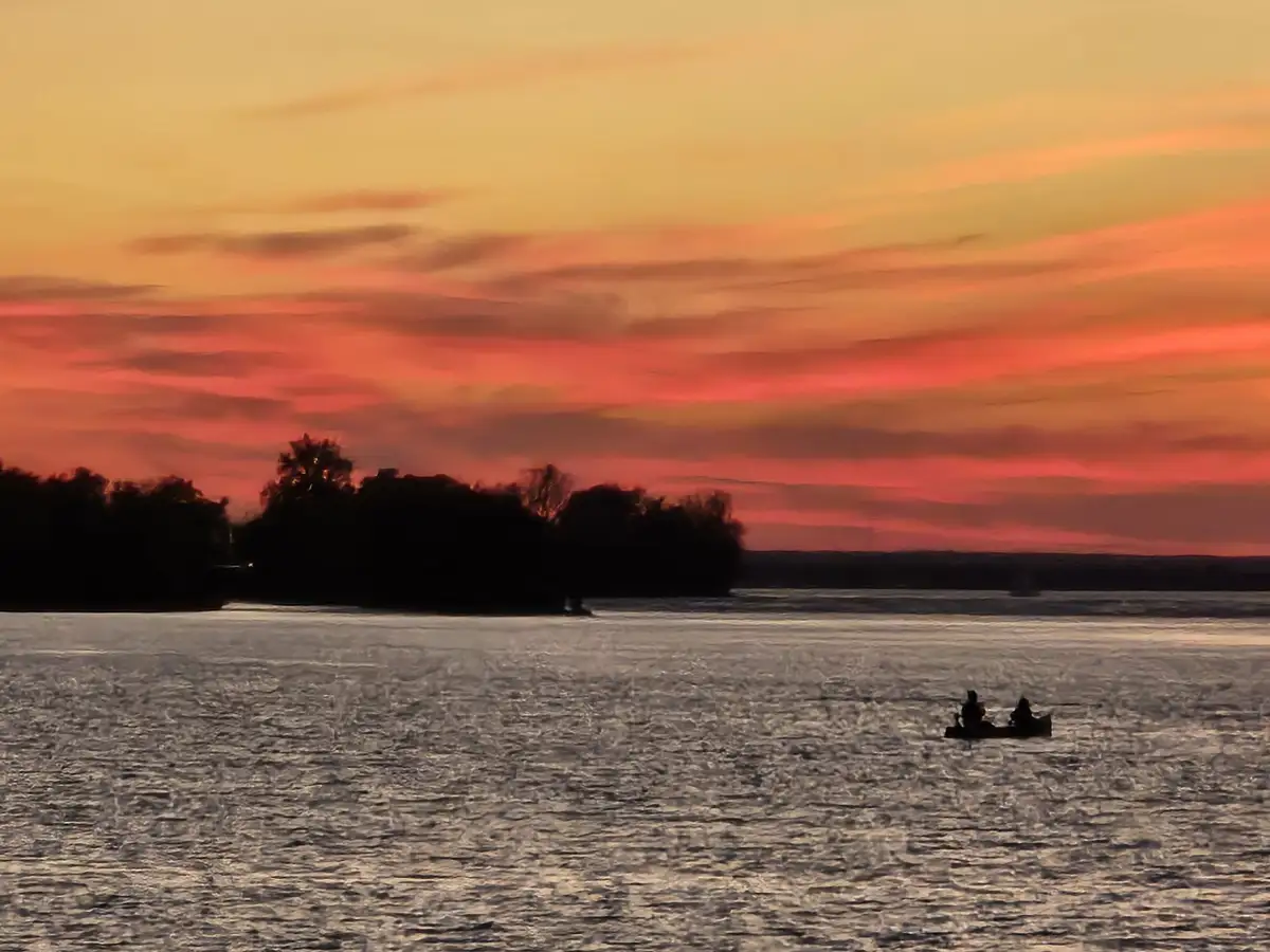 A serene sunset over a body of water with silhouettes of trees on the horizon and two people in a small boat. The sky is…