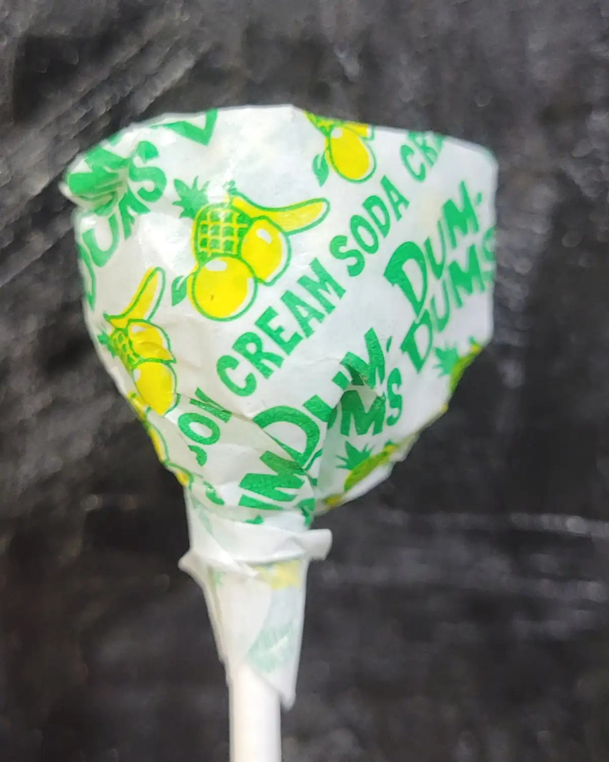 A close-up of a crumpled white wrapper with green and yellow text and pineapple graphics, identifying the candy as 'DUM DUMS…
