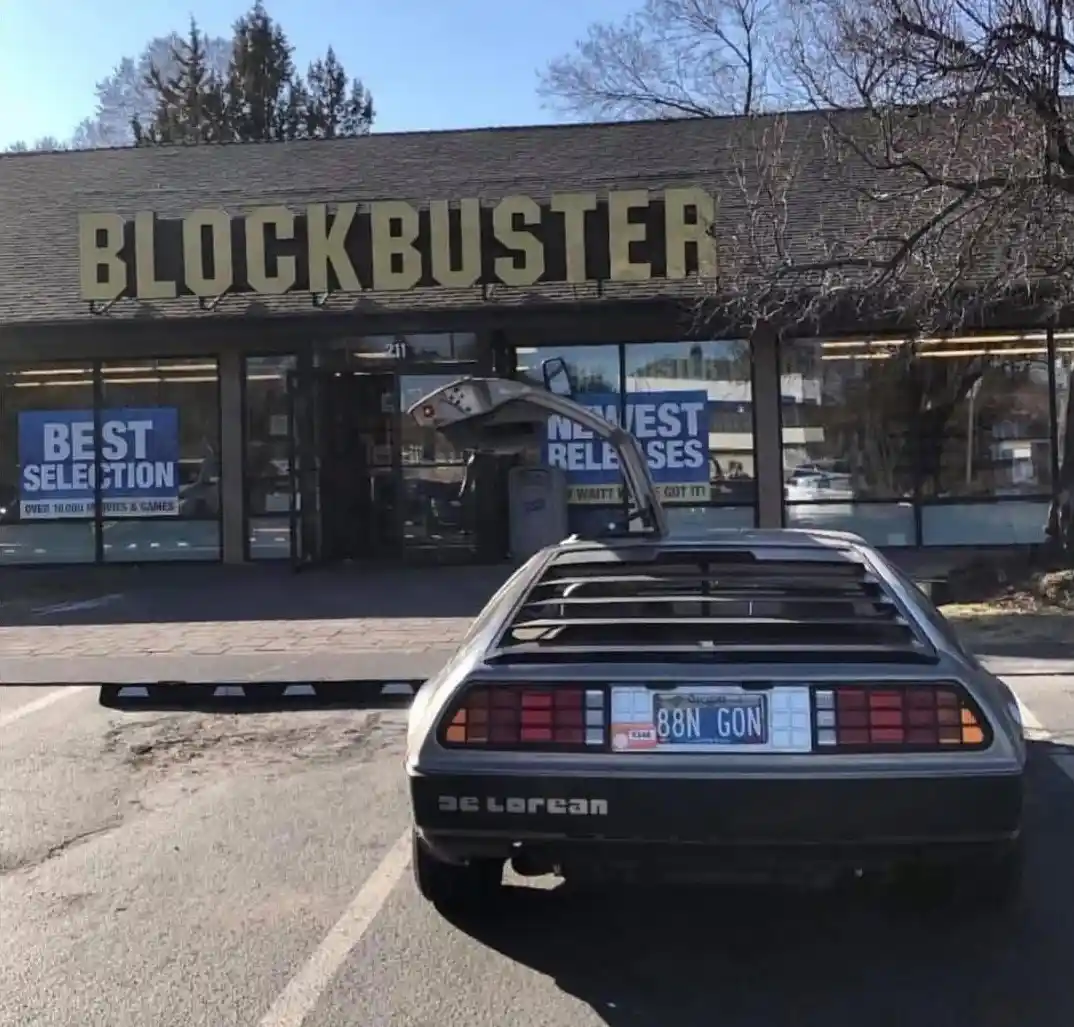 A silver DeLorean time machine is parked in front of a closed Blockbuster video store, its gull-wing door open. The car's…