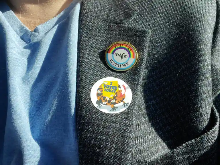 A close-up of a person's chest wearing a grey sweater over a blue shirt, displaying two pins: one circular pin with a…
