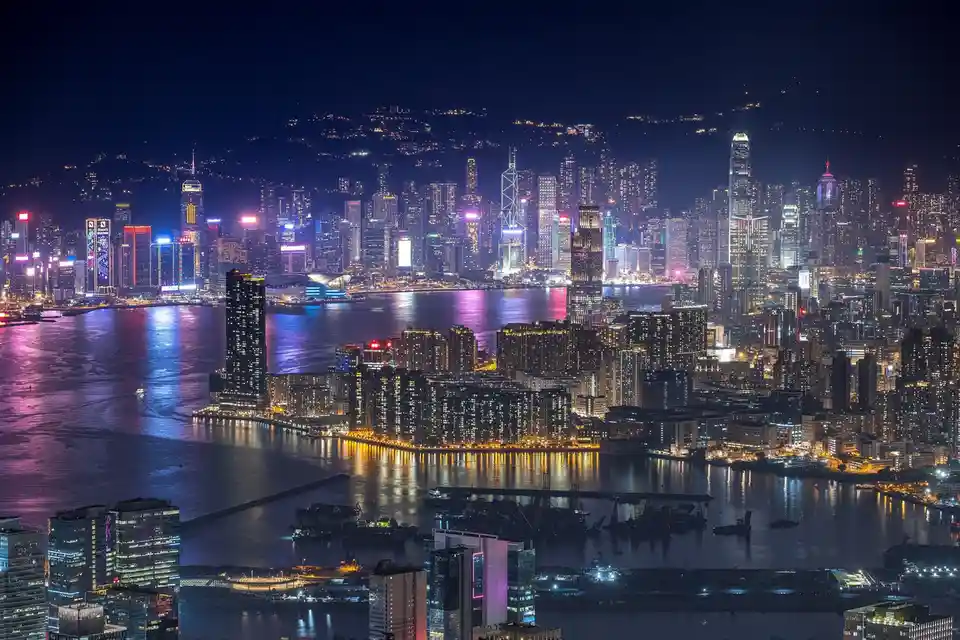 A vibrant nighttime cityscape of Hong Kong, featuring a glittering skyline across Victoria Harbour with illuminated…
