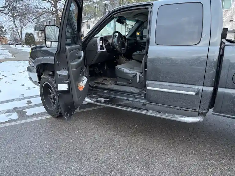 A gray pickup truck is parked in a snowy lot with its driver's side door open, revealing a worn interior. The vehicle…