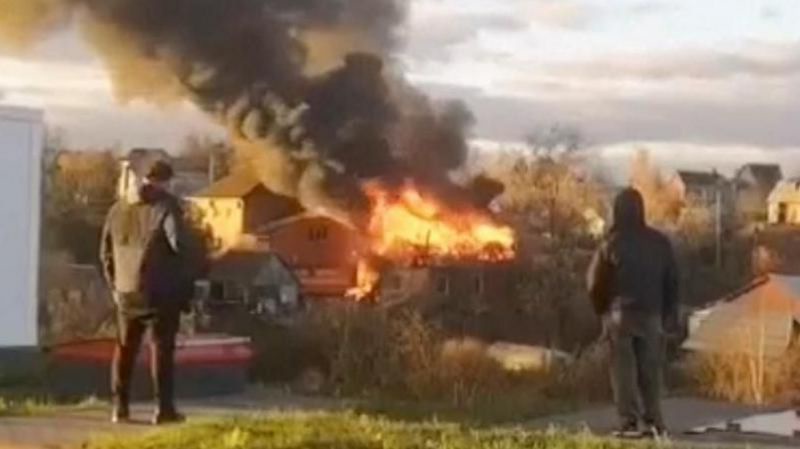 Two people stand in a grassy area watching a large house burn with bright orange flames and thick black smoke billowing into…