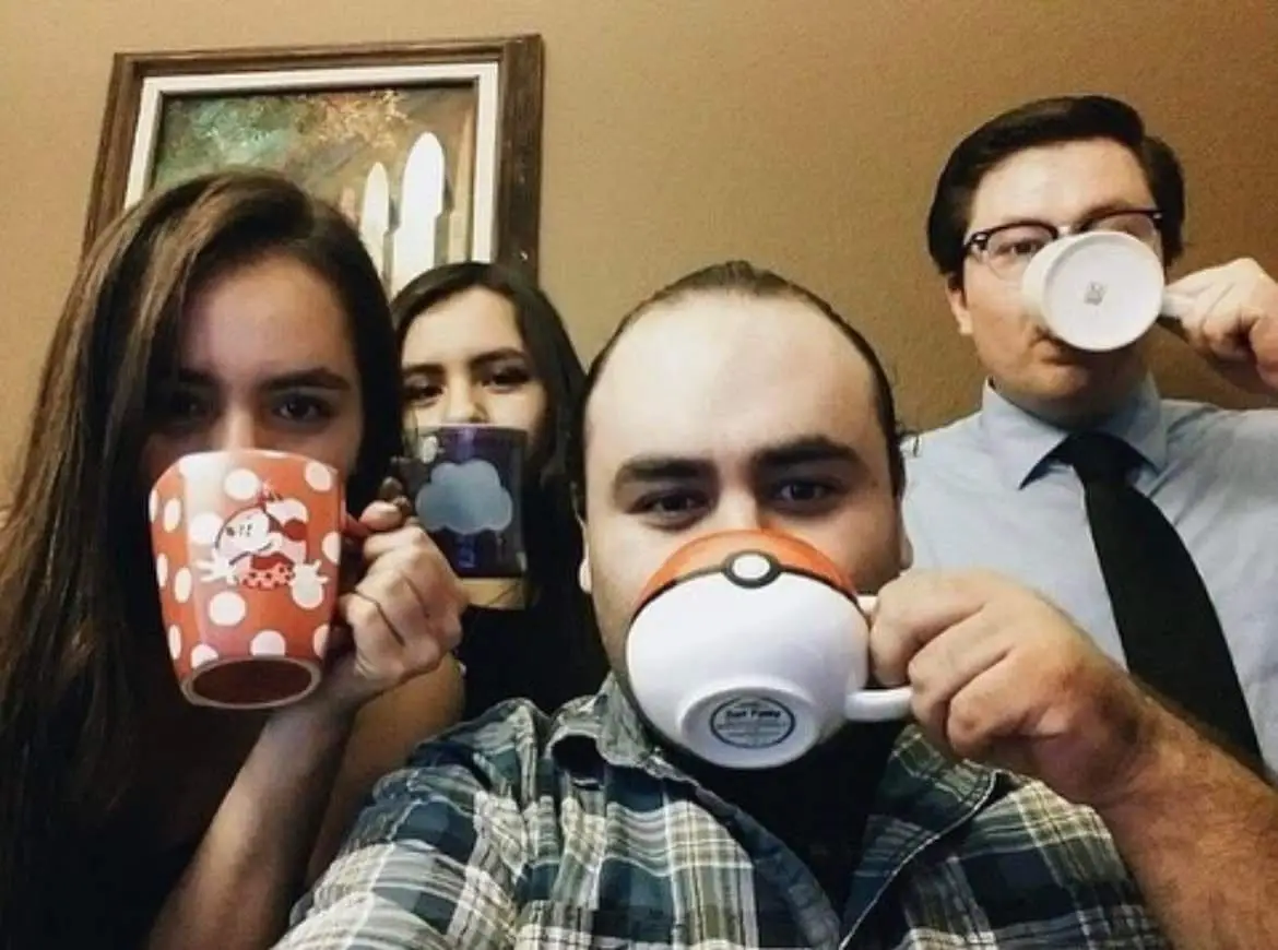 Four people in a selfie pose, each holding a mug up to their face as if drinking, but all are looking directly at the camera…