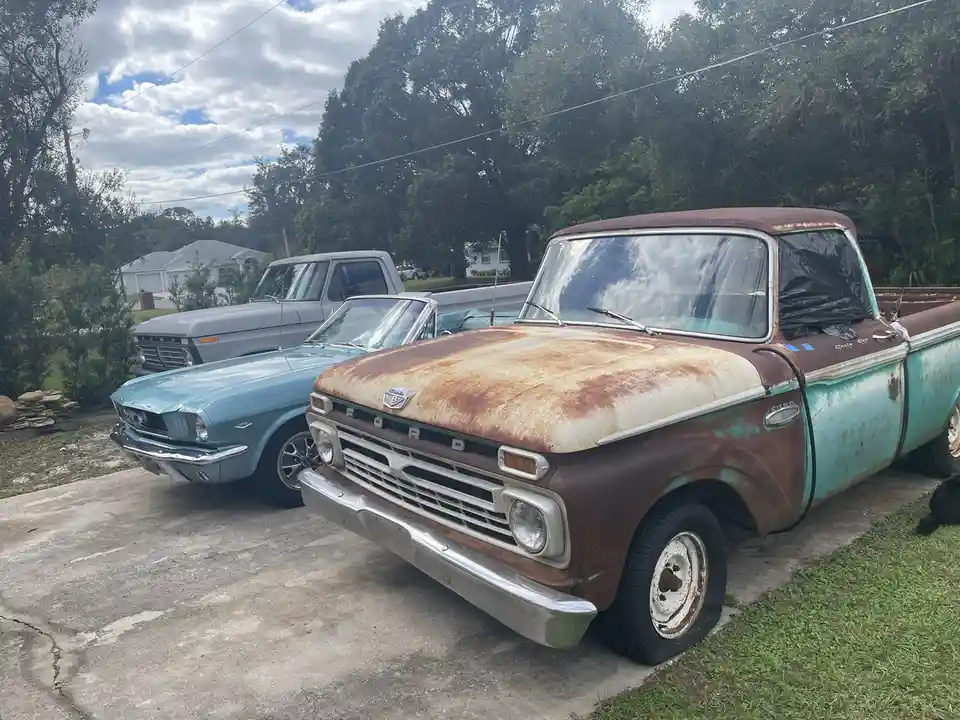 A rusty, weathered vintage Ford pickup truck with peeling turquoise paint and a rusted hood is parked in a driveway…