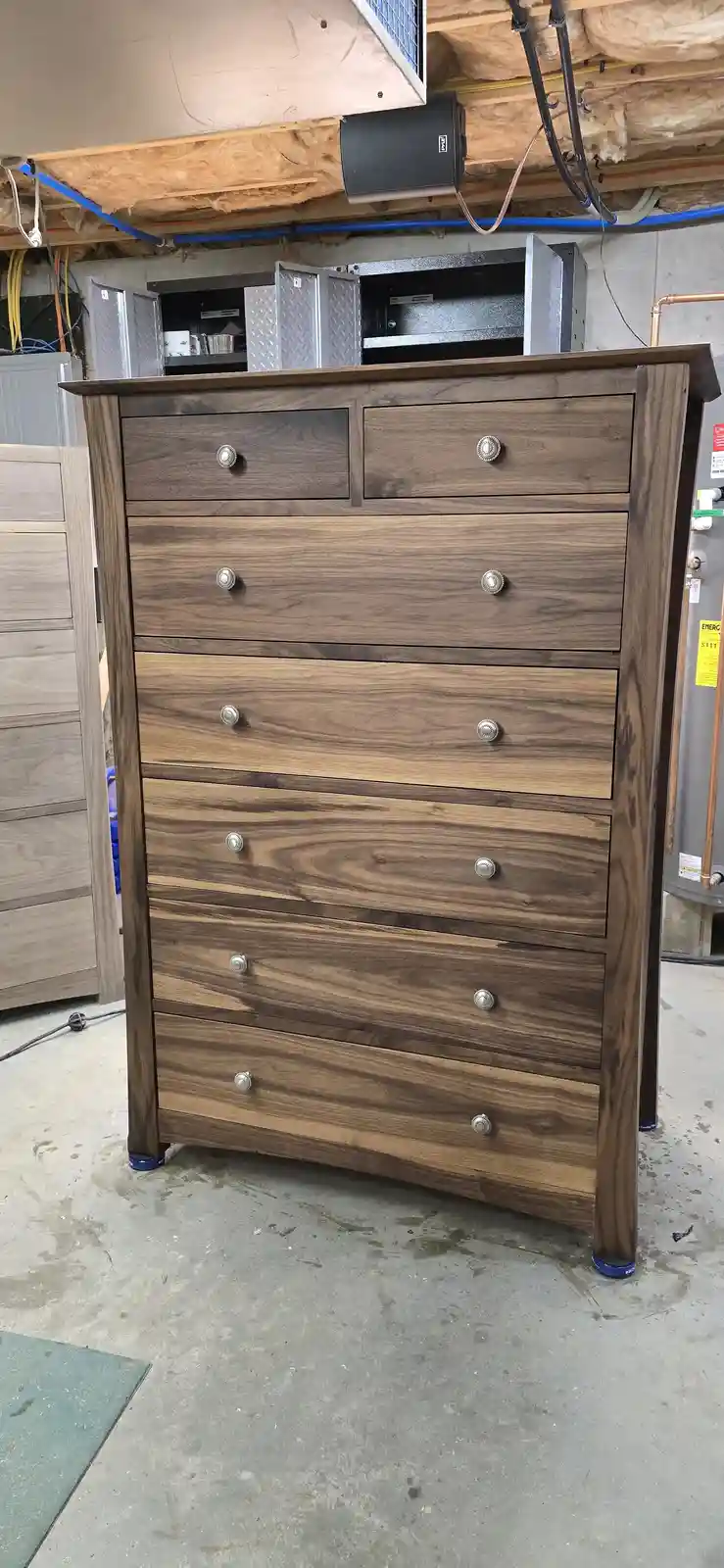 A tall wooden dresser with multiple drawers and silver knobs stands in a cluttered basement or workshop, visibly leaning at…