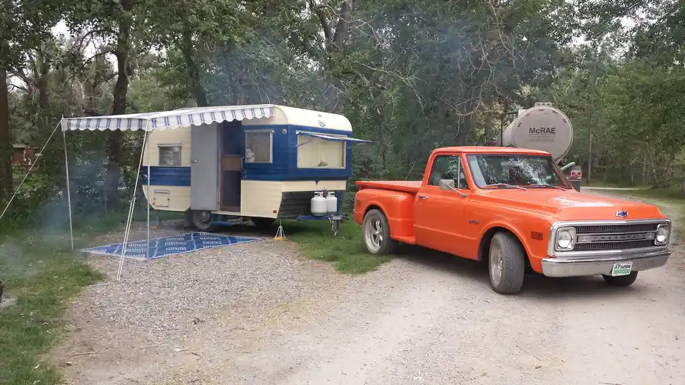 An orange 1960s Chevrolet C10 pickup truck is parked on a gravel lot next to a vintage blue and cream camper trailer with…
