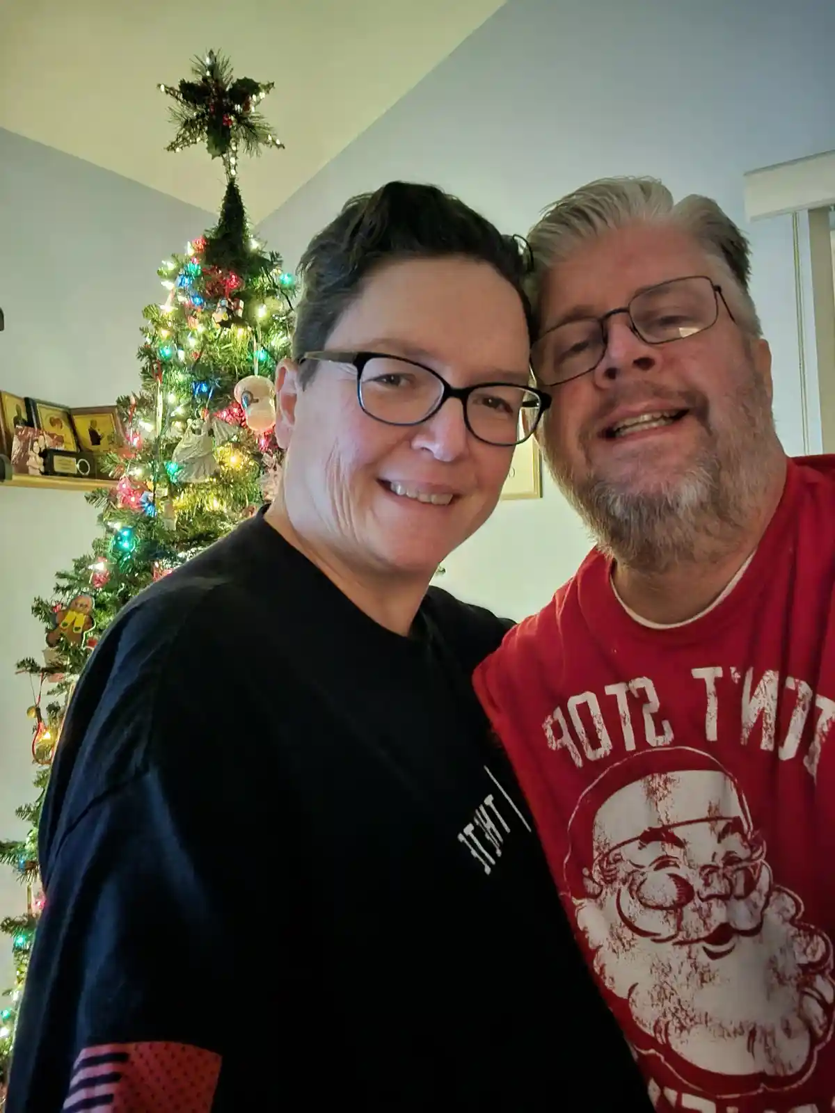 Two smiling people pose for a selfie in front of a decorated Christmas tree with colorful lights and ornaments. The person…