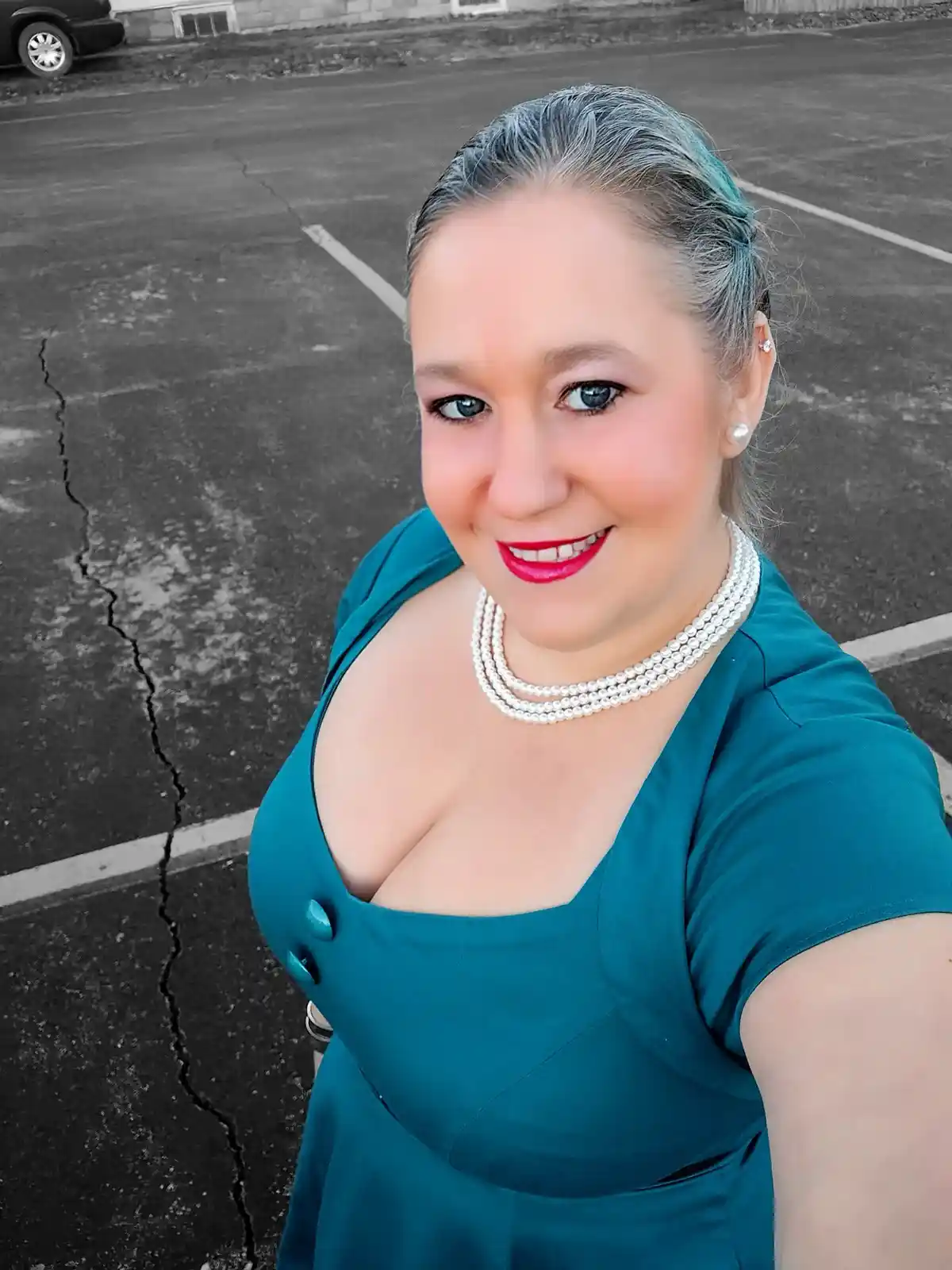 A woman in a teal dress with pearl necklace and bright red lipstick smiles at the camera in a parking lot, with the…
