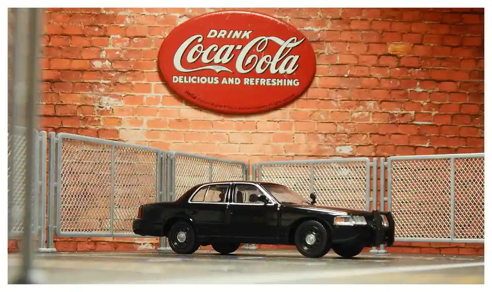 A black police car model is parked in front of a chain-link fence, with a large red oval Coca-Cola sign reading 'DRINK…