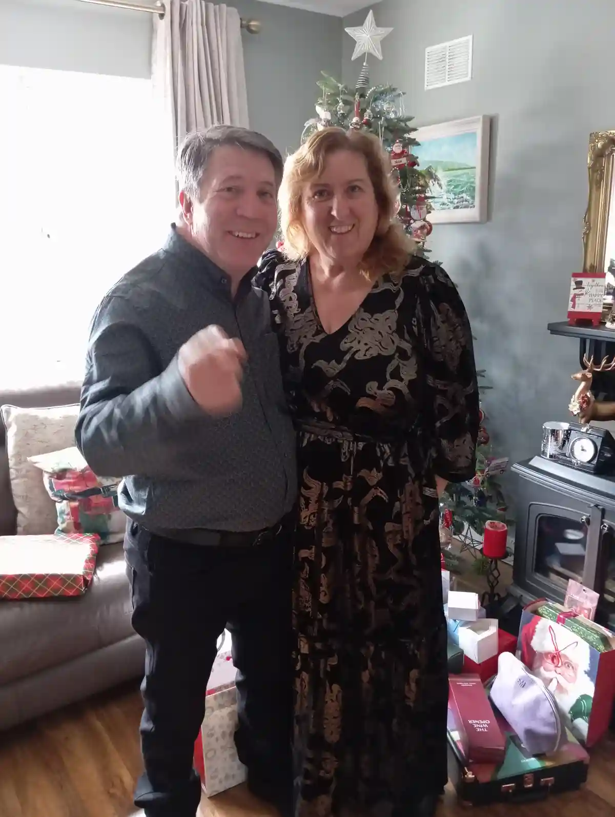 A man and woman stand smiling in a living room decorated for Christmas, with a tree, gifts, and a fireplace visible. The man…