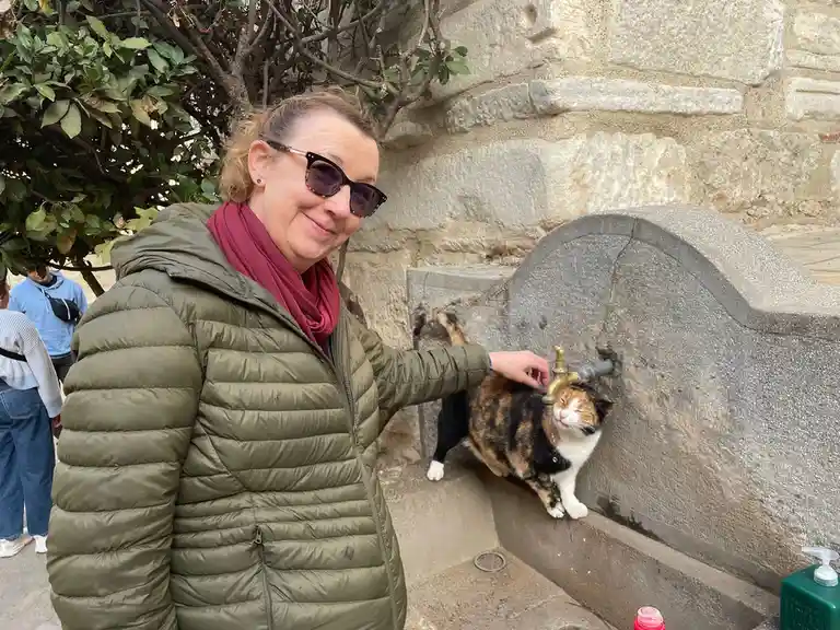A woman in a green puffer jacket and sunglasses smiles while her hand rests on a calico cat that is drinking from a stone…