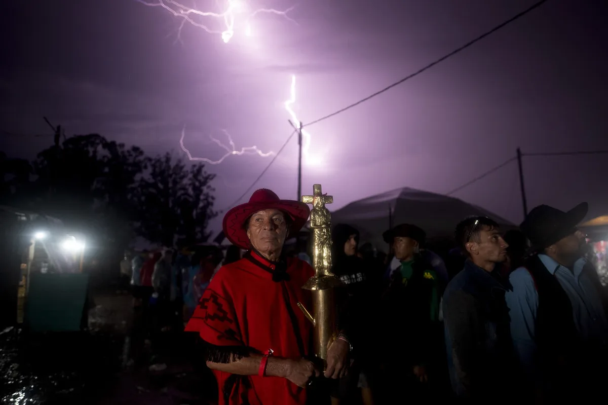 A man in a red poncho and wide-brimmed hat stands solemnly holding a golden religious statue under a stormy sky with vivid…
