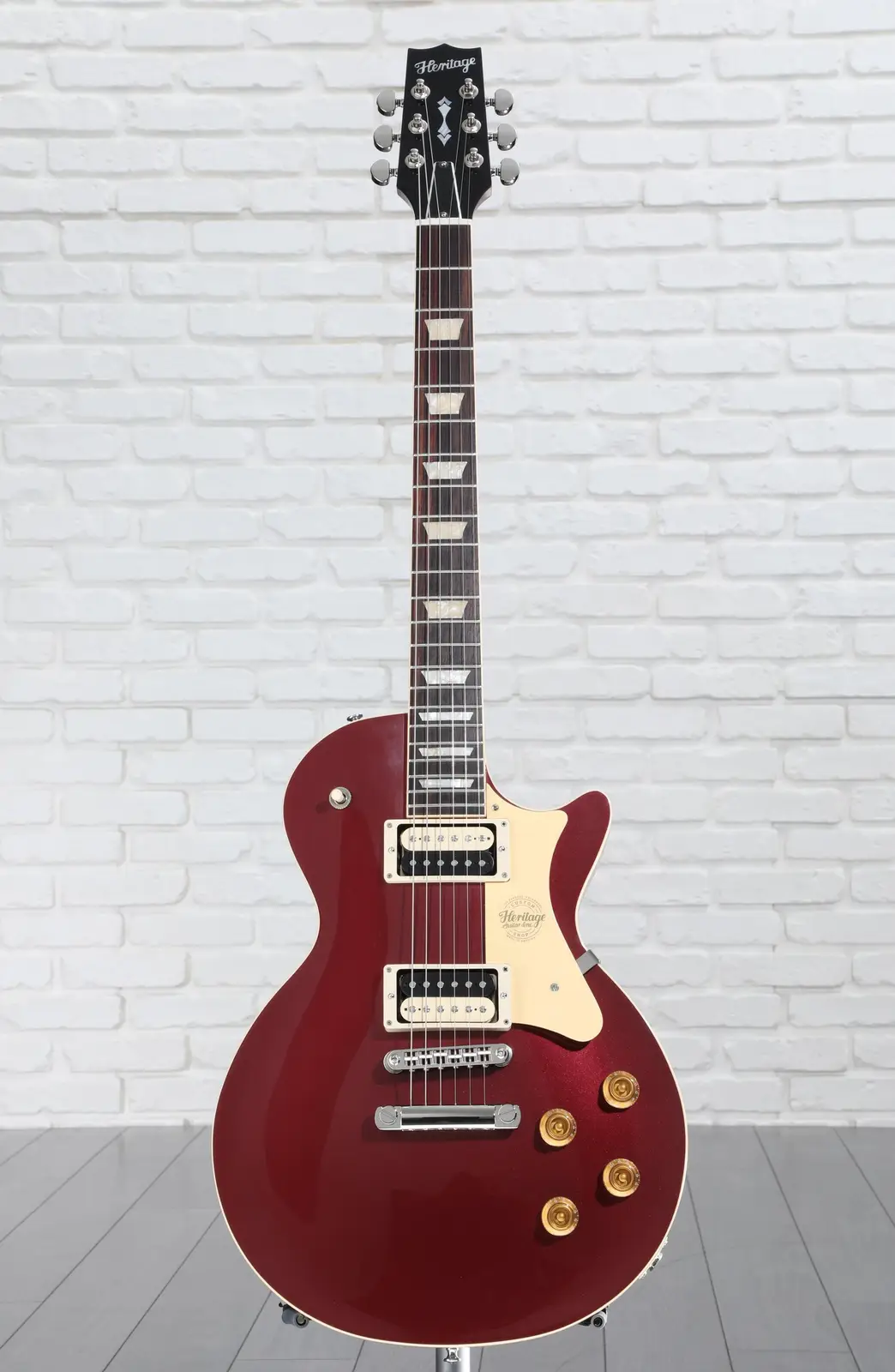 A red Heritage electric guitar with a white pickguard and gold hardware stands upright against a white brick wall. The…