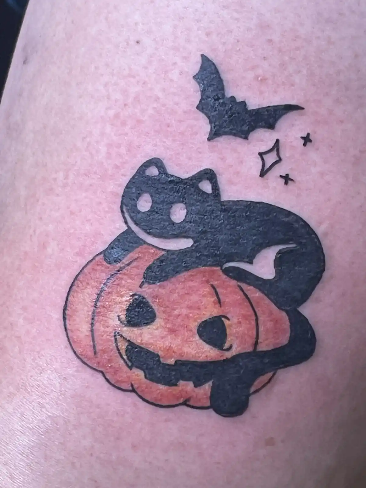 A freshly inked tattoo on skin shows a cartoon-style black cat lounging atop a smiling orange jack-o'-lantern with carved…