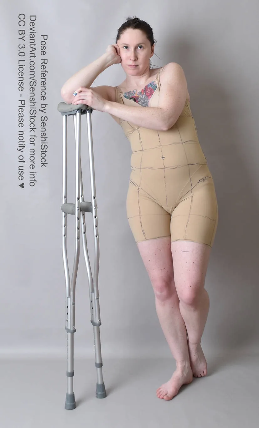 A woman with a colorful tattoo on her chest stands barefoot next to a pair of crutches, wearing a beige form-fitting…