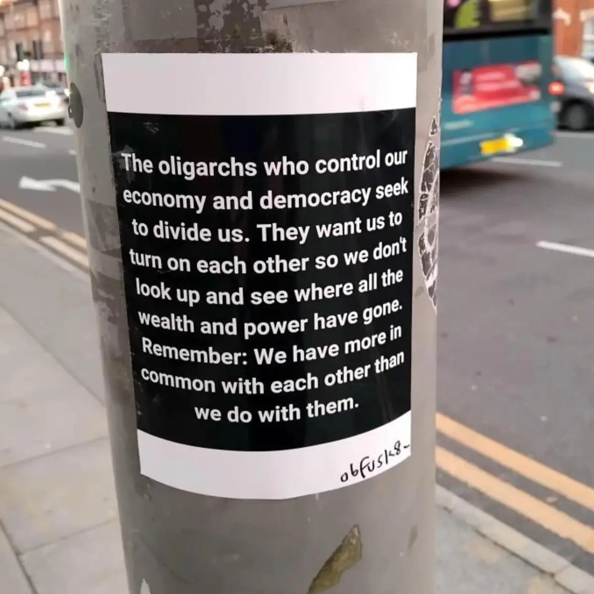 A black-and-white sticker is affixed to a weathered metal pole on a city sidewalk, with cars and a bus visible in the…