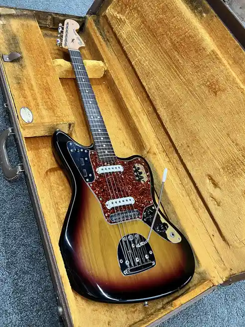 A sunburst Fender Jaguar electric guitar rests inside its worn, yellow-felt-lined hardshell case, with a small white…