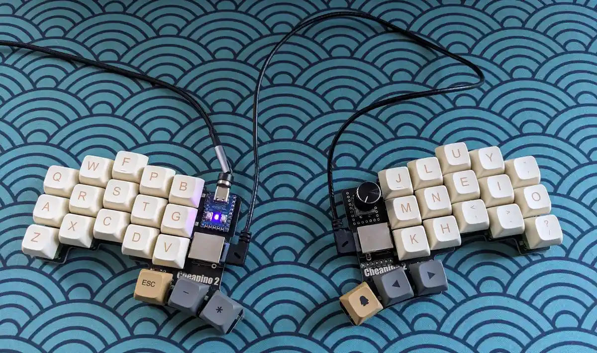 Two split ergonomic keyboard halves labeled 'Cheapino 2' sit on a blue wave-patterned surface, connected by black cables…