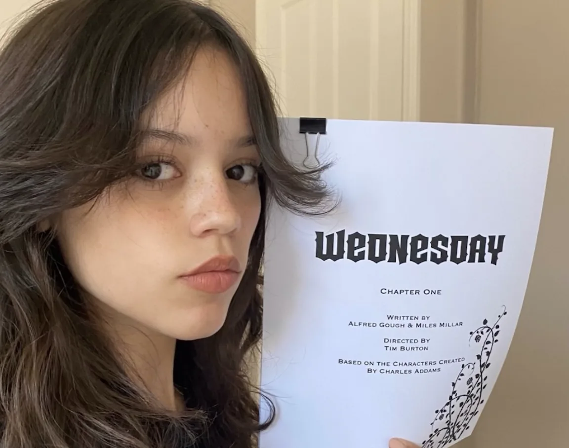 A young woman with dark hair and freckles holds up a script titled 'WEDNESDAY' with 'CHAPTER ONE' and credits for Alfred…