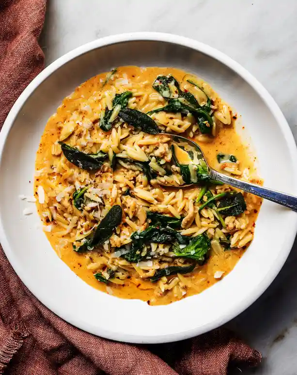 A top-down photo of a white bowl filled with creamy orzo pasta mixed with spinach, sage leaves, and ground meat, garnished…