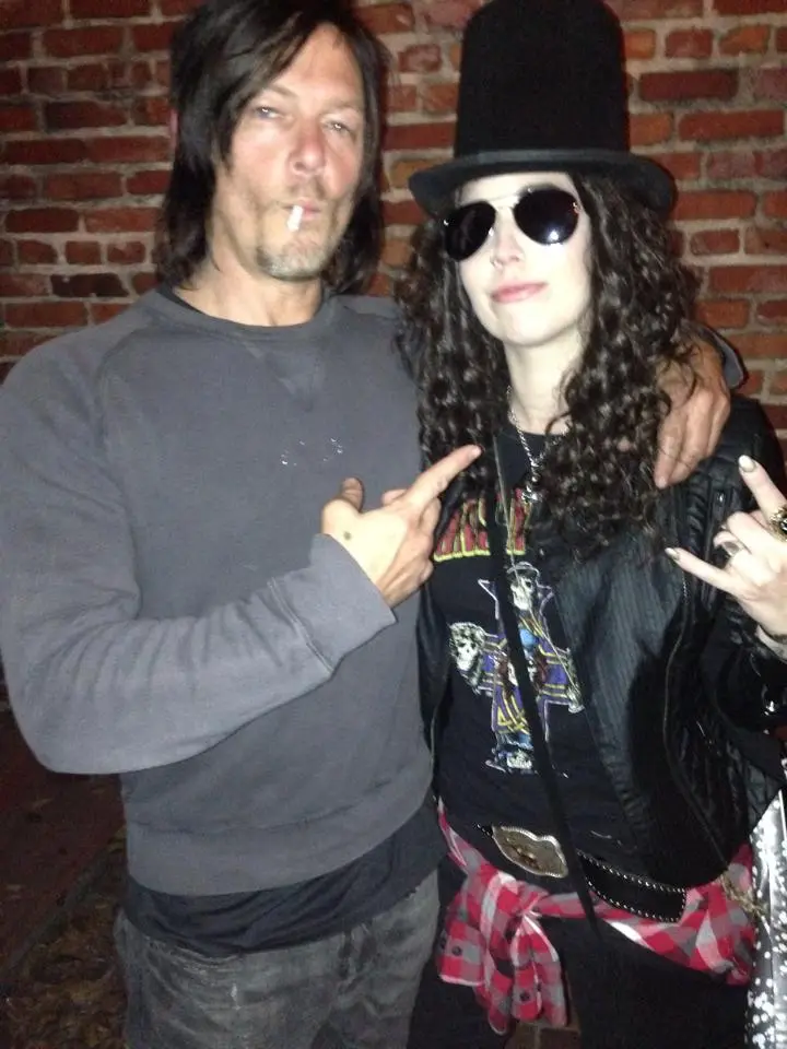 A man with long dark hair and a cigarette in his mouth, wearing a grey sweatshirt, points at a woman beside him. The woman…