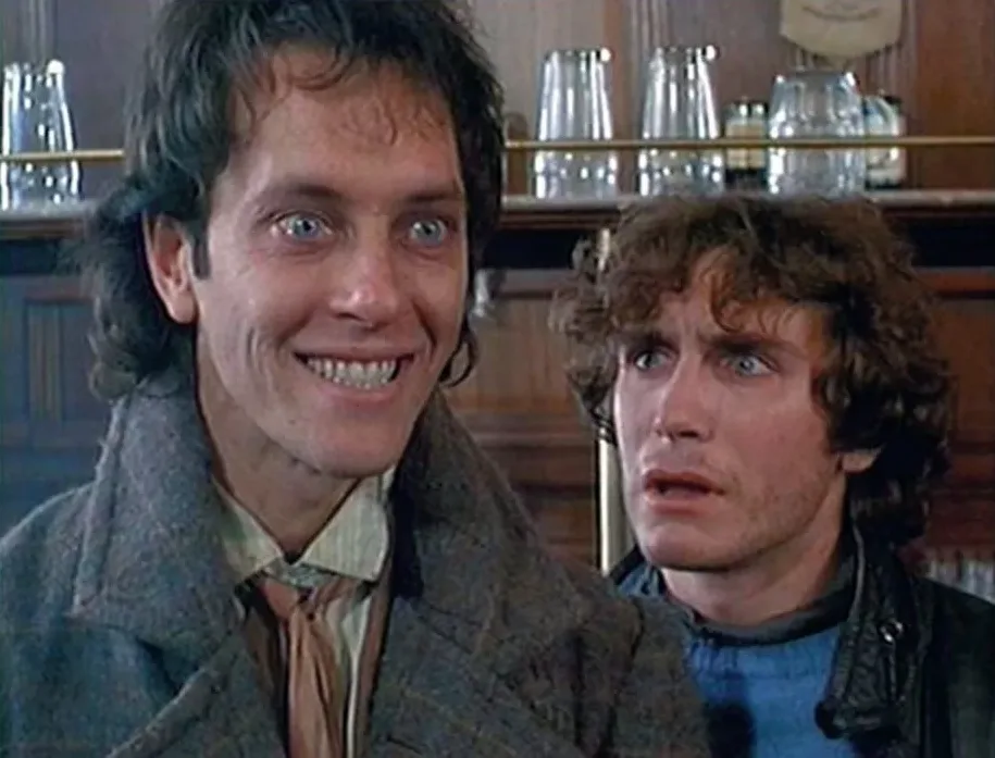 Two men are shown in a close-up shot inside what appears to be a bar or pub, with shelves of glassware behind them. The man…