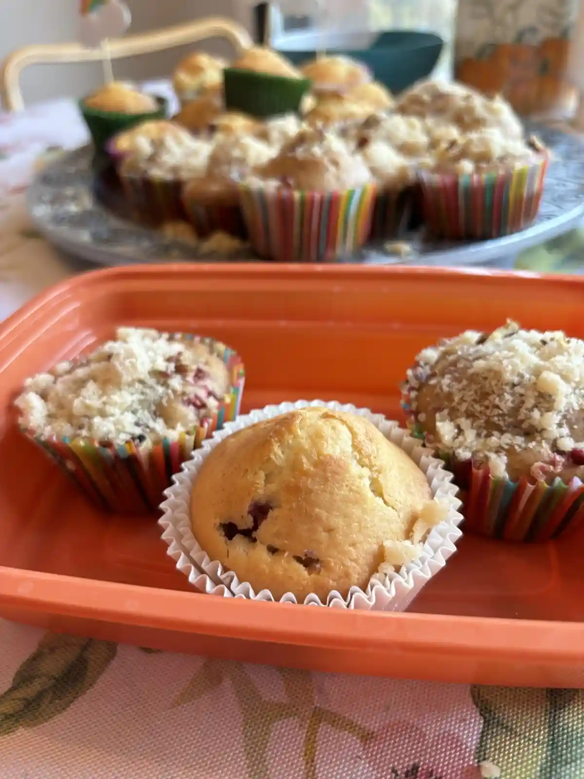 A close-up photo shows three muffins on an orange plastic tray in the foreground, with a plate of more muffins blurred in…