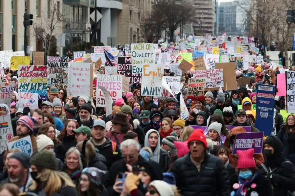 A large crowd of protesters marches down a city street, holding numerous handmade signs with slogans about reproductive…