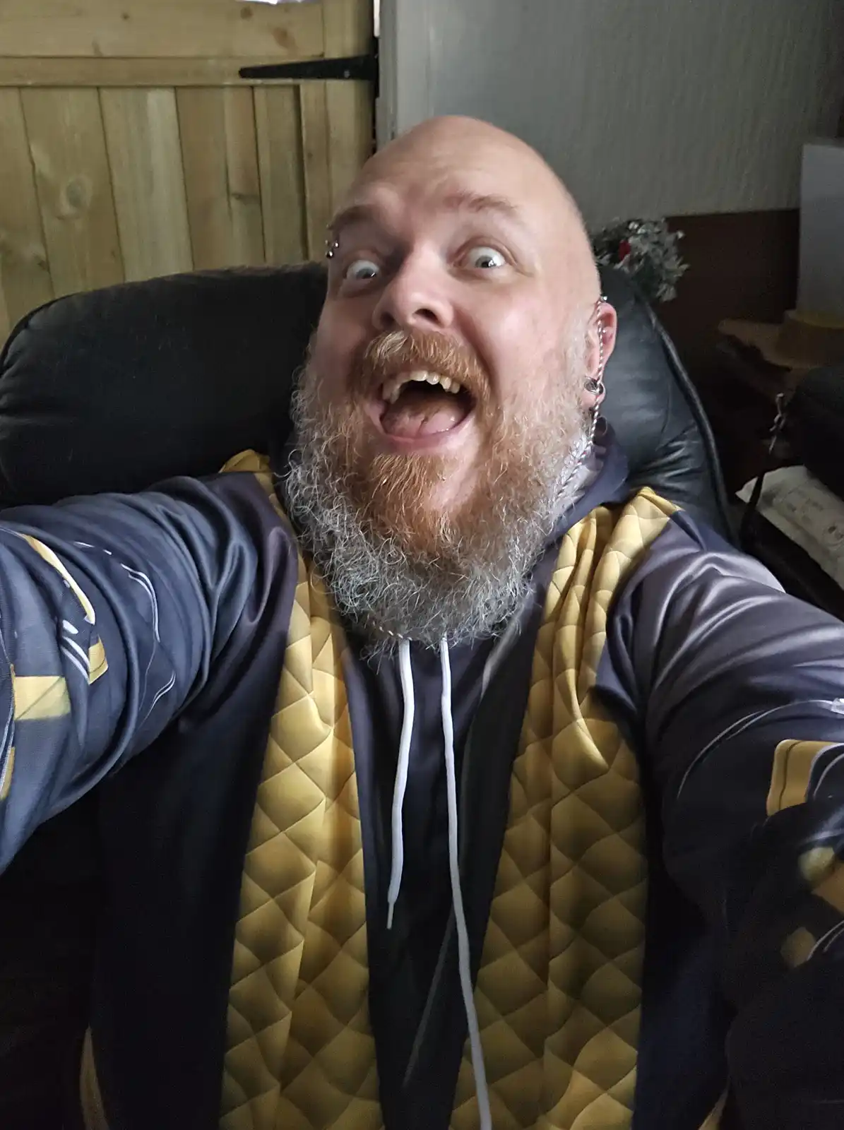 A bald man with a long, graying beard and multiple ear piercings takes a selfie while sitting in a black chair. He has a…