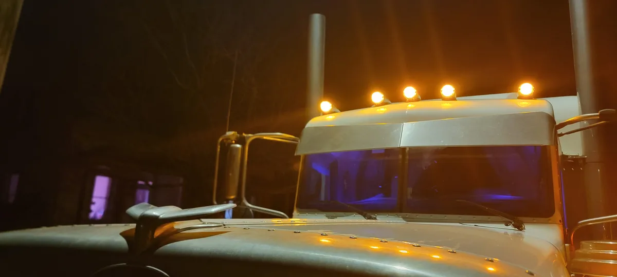 A close-up nighttime photo of a white semi-truck's cab, featuring its illuminated amber roof lights and hood lights against…