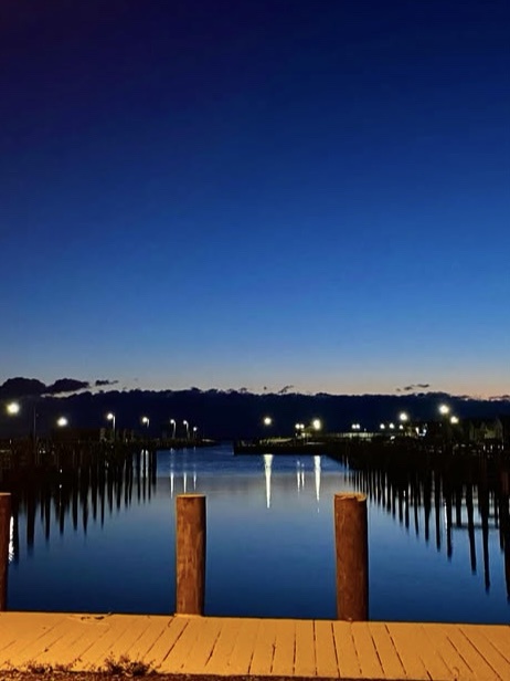 A serene twilight photo of a calm waterfront with wooden pilings and a dock, streetlights reflecting in the water under a…