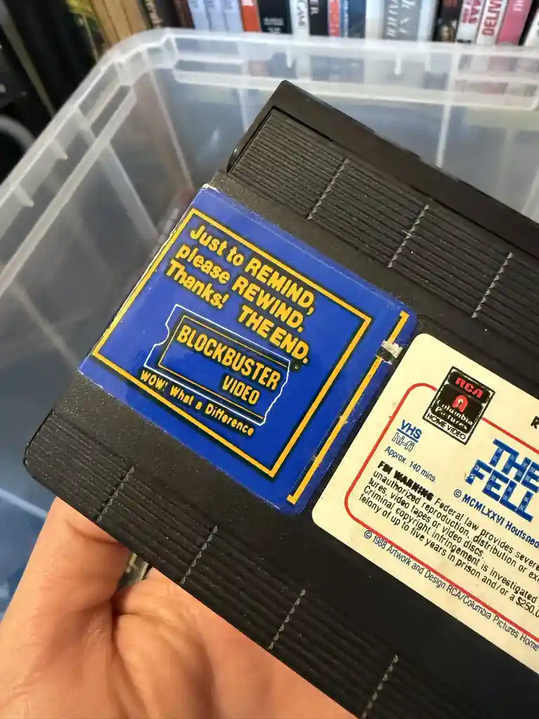 A hand holds a VHS tape with a Blockbuster Video sticker that reads 'Just to REMIND, please REWIND. Thanks! THE END.' The…