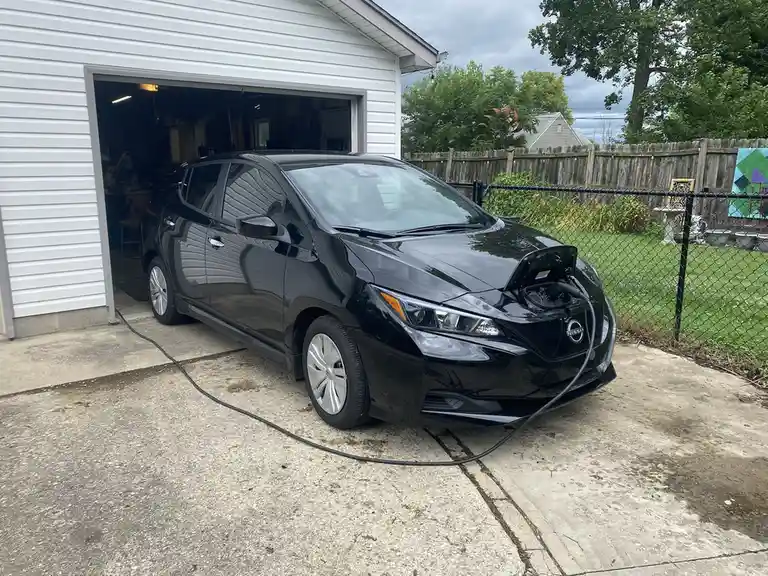 A black Nissan Leaf electric car is parked on a concrete driveway in front of a white garage, plugged into a charging…