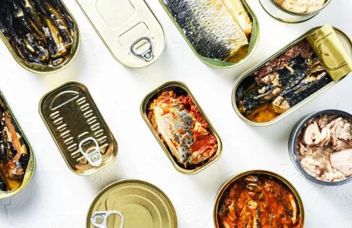An overhead shot of numerous open and closed canned fish products scattered across a white surface. The cans contain various…