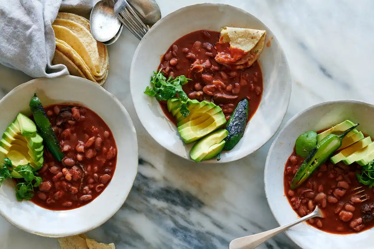 Three bowls of red bean stew topped with sliced avocado, cilantro, and charred green chiles sit on a marble surface. One…