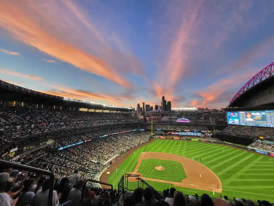 A wide-angle photo of a baseball stadium during sunset, showing a packed crowd, a green field with players, and a city…