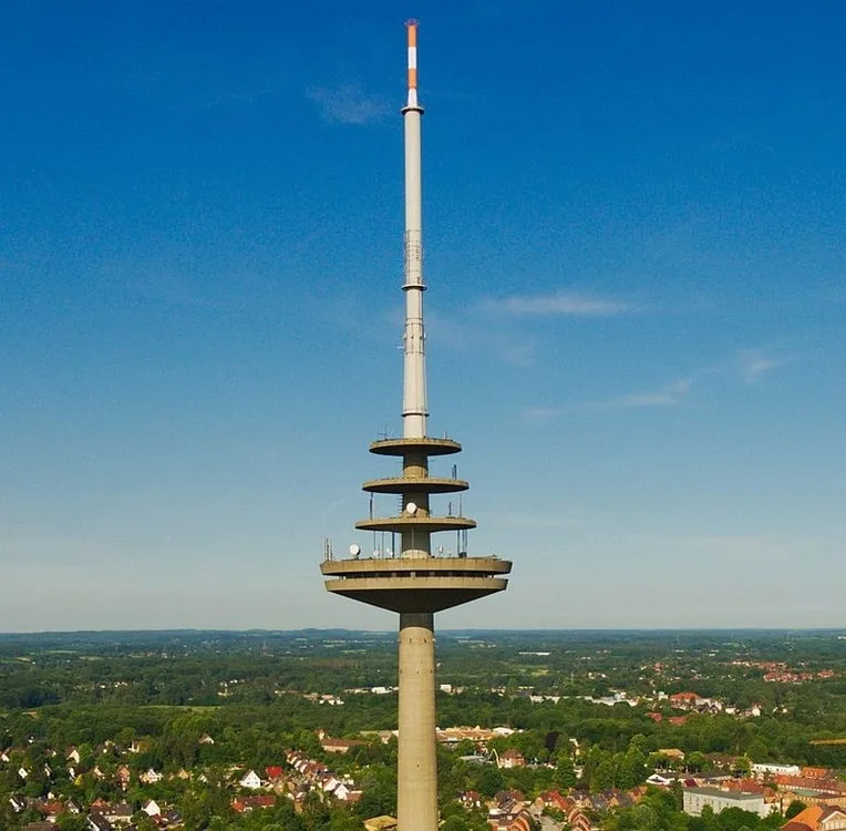 An aerial photo shows a tall, slender concrete telecommunications tower with multiple circular platforms and antennas, set…