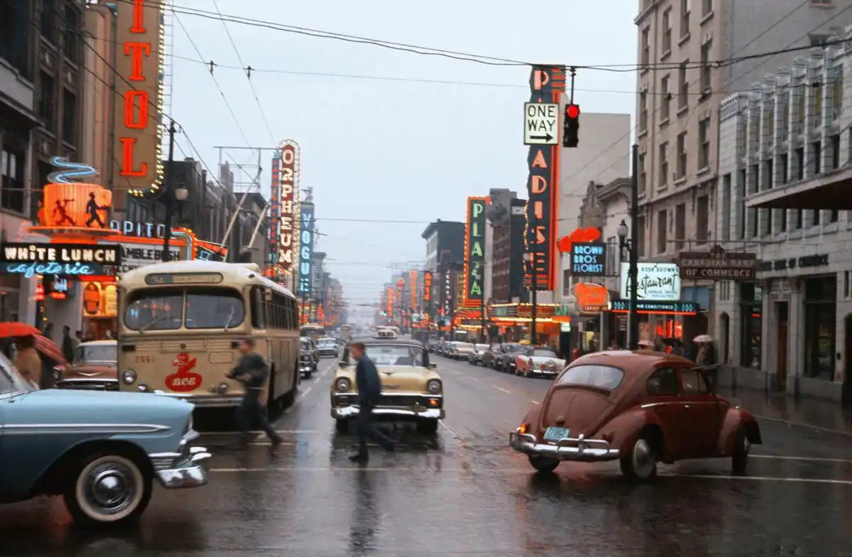 A vintage photograph of a rainy, bustling city street lined with classic cars and bright neon signs for theaters like the…