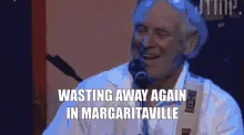 A still image of an older man with white hair, wearing a white shirt and tie, singing into a microphone on stage. The text…