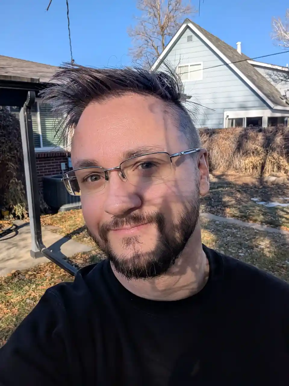 A man with glasses and a beard smiles at the camera outdoors in front of a house with autumn leaves on the ground. The…