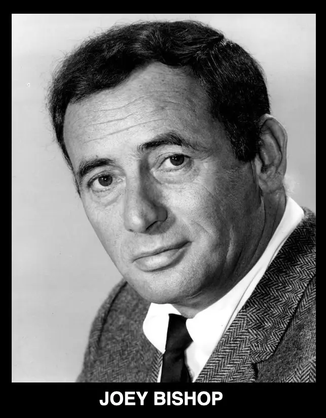 A black-and-white portrait of a man with dark hair, wearing a herringbone-patterned suit jacket, white shirt, and dark tie…
