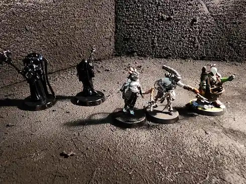 Five painted miniature figurines stand in a line on a textured gray surface against a rough wall. The two figures on the…