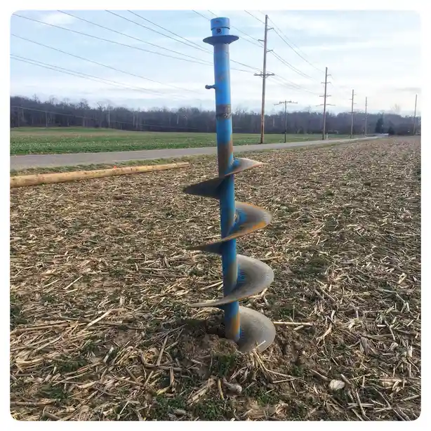 A blue, rusted auger drill bit stands partially embedded in a tilled field covered in crop residue, with a dirt path and…