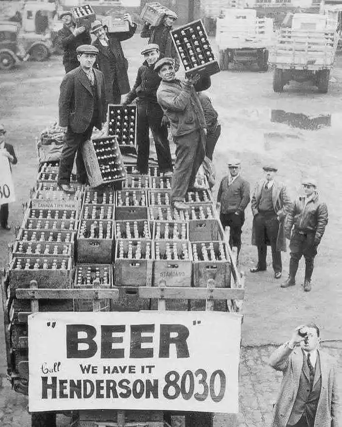A black-and-white historical photo shows men loading crates of beer bottles onto a truck, with a large banner reading 'BEER…