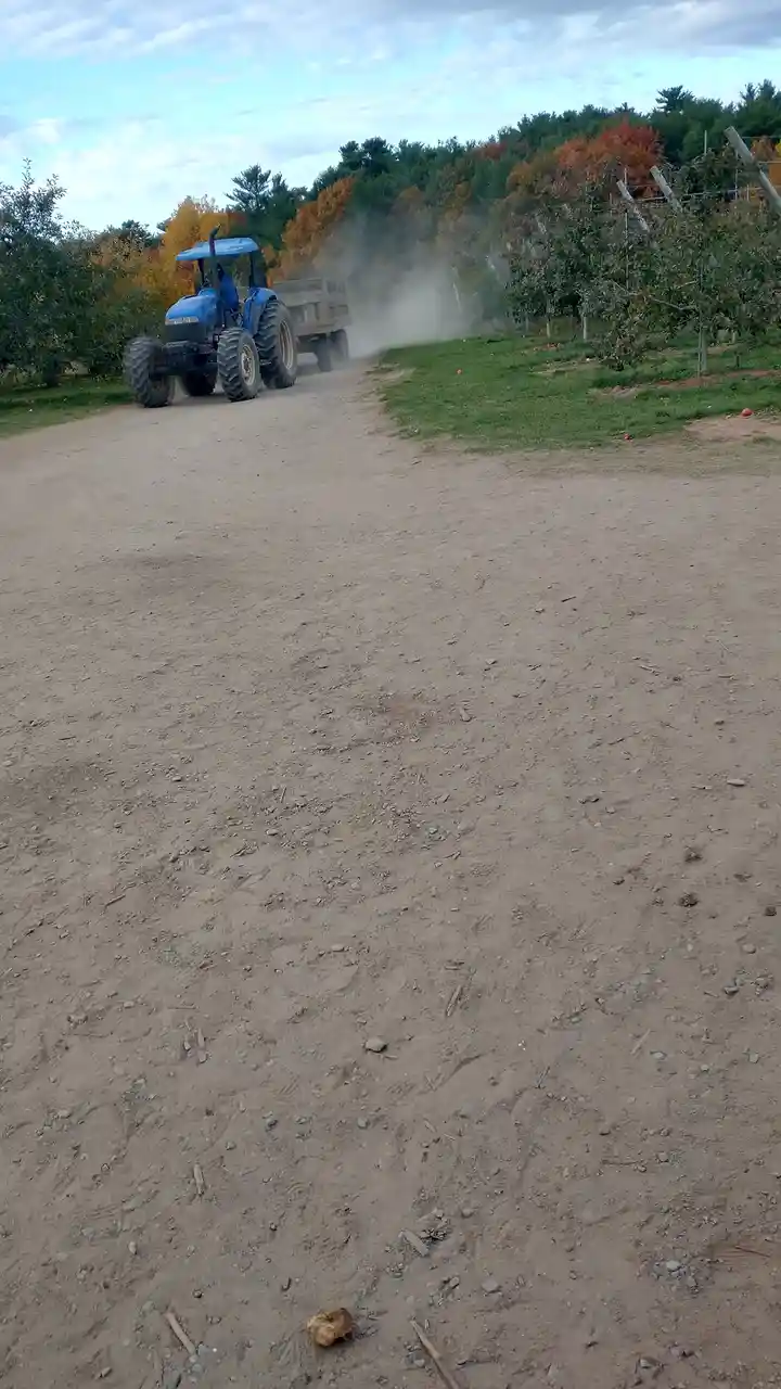 A blue tractor pulling a trailer kicks up dust on a dirt road in an orchard with autumn-colored trees. The overlaid text…