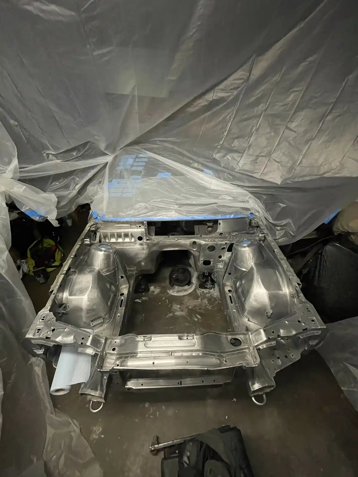 A stripped, silver-painted car chassis sits in a garage, surrounded by plastic sheeting and blue painter's tape. The text…