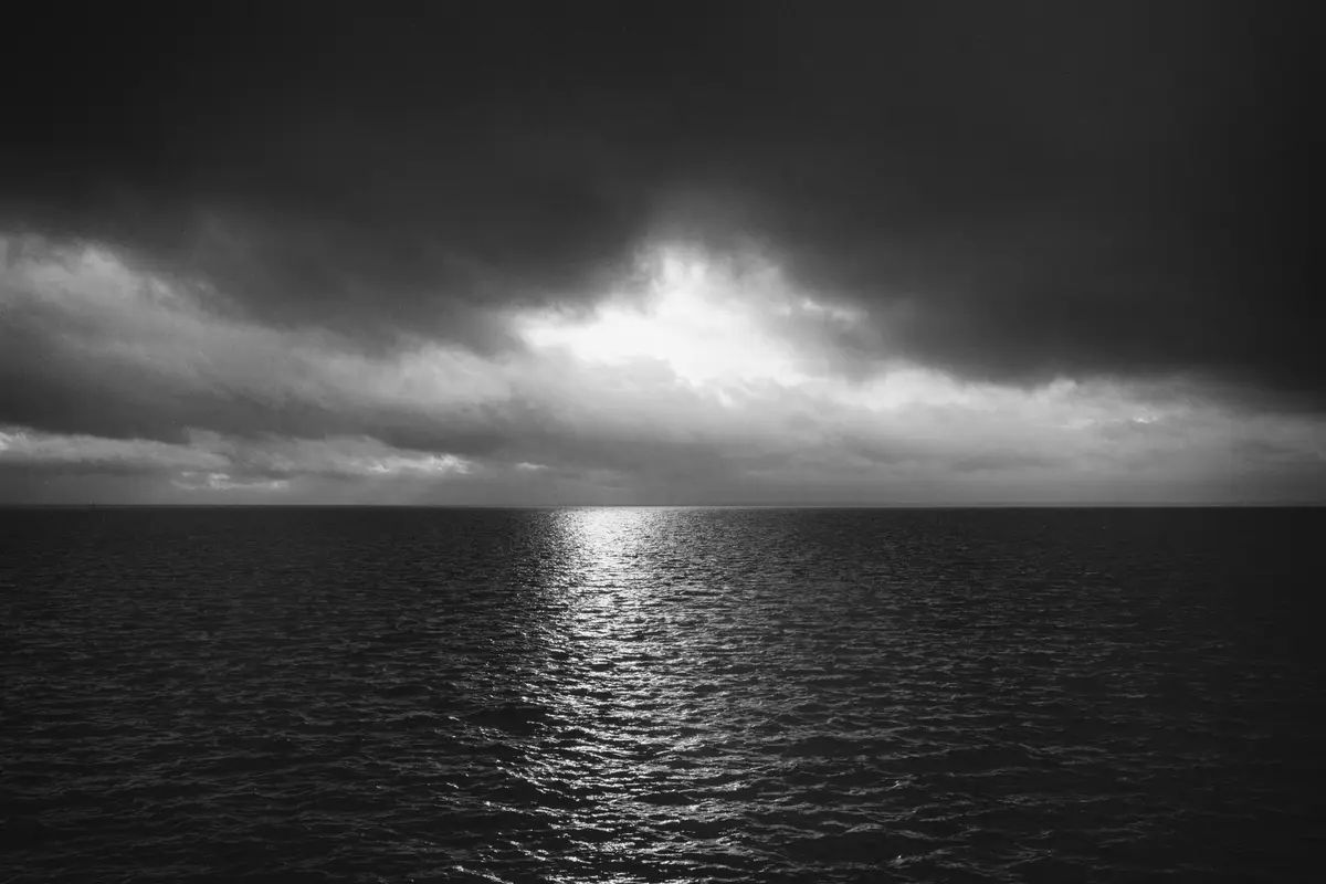 A black and white photograph shows a vast, dark ocean under a heavy, stormy sky with a bright patch of sunlight breaking…